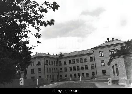 Sloviansk, 1956-1960, via Shevchenko. Questa storica fotografia d'archivio mostra la robusta architettura in mattoni a più piani dell'ospedale Lenin. Diversi individui, probabilmente personale medico e visitatori, sono riuniti vicino all'ingresso principale sotto l'ombra di alberi maturi. Le motociclette d'epoca con sidecar sono parcheggiate ordinatamente lungo il vialetto asfaltato. L'edificio presenta numerose grandi finestre, tetti piastrellati ed elementi strutturali classici che mostrano il sistema sanitario del tranquillo Donbas. Foto Stock