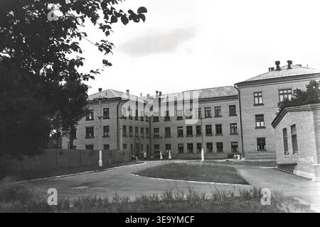 Sloviansk, 1956-1960, via Shevchenko. Questa storica fotografia d'archivio mostra la robusta architettura in mattoni a più piani dell'ospedale Lenin. Diversi individui, probabilmente personale medico e visitatori, sono riuniti vicino all'ingresso principale sotto l'ombra di alberi maturi. Le motociclette d'epoca con sidecar sono parcheggiate ordinatamente lungo il vialetto asfaltato. L'edificio presenta numerose grandi finestre, tetti piastrellati ed elementi strutturali classici che mostrano il sistema sanitario del tranquillo Donbas. Foto Stock