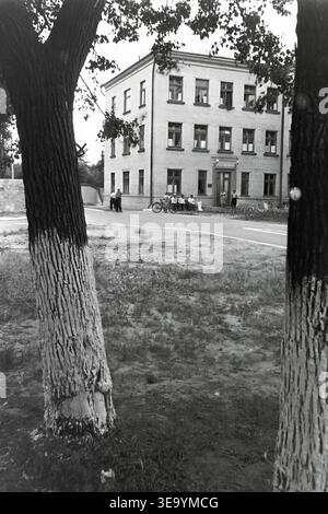Sloviansk, 1956-1960, via Shevchenko. Questa storica fotografia d'archivio mostra la robusta architettura in mattoni a più piani dell'ospedale Lenin. Diversi individui, probabilmente personale medico e visitatori, sono riuniti vicino all'ingresso principale sotto l'ombra di alberi maturi. Le motociclette d'epoca con sidecar sono parcheggiate ordinatamente lungo il vialetto asfaltato. L'edificio presenta numerose grandi finestre, tetti piastrellati ed elementi strutturali classici che mostrano il sistema sanitario del tranquillo Donbas. Foto Stock
