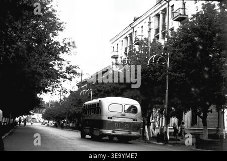 Sloviansk, 1956-1960, via Shevchenko. Questa splendida fotografia d'archivio storico cattura il fascino di un vivace viale urbano. Un classico autobus cittadino ZIS-155 con finestrini posteriori ben visibili sta percorrendo l'ampia strada, passando sotto la tettoia di alberi lussureggianti e maturi. Eleganti lampioni lungo il marciapiede, completano i balconi ornati e la facciata monumentale dell'edificio residenziale a più piani sulla destra. Un'istantanea di trasporti dinamici e architettura elegante nella tranquilla Donbas. Foto Stock