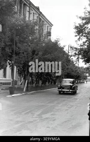 Sloviansk, 1956-1960, via Shevchenko. Questa splendida fotografia d'archivio storico cattura il grande fascino di un vivace viale urbano. Un'elegante e classica autovettura di Pobeda guida attivamente lungo l'ampia strada asfaltata, passando sotto la tettoia di alberi lussureggianti e maturi. Eleganti lampioni ad arco fiancheggiano il marciapiede, completando i balconi ornati e la facciata monumentale dell'edificio residenziale a più piani sulla destra. Un'istantanea di trasporti dinamici e architettura elegante nella tranquilla Donbas. Foto Stock