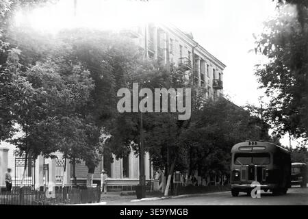 Sloviansk, 1956-1960, via Shevchenko. Questa affascinante fotografia d'archivio storica offre una meravigliosa prospettiva del transito urbano quotidiano. Un classico autobus cittadino ZIS-155, con la sua caratteristica carrozzeria arrotondata, percorre l'ampia strada asfaltata. Un pedone in una tuta bianca cammina lungo il marciapiede, circondato da alberi ombreggiati splendidamente paesaggistici ed eleganti lampioni ad arco. Dietro il fitto fogliame, la solida facciata di un grande edificio residenziale a più piani si erge alto, riflettendo la pace di Donbas. Foto Stock