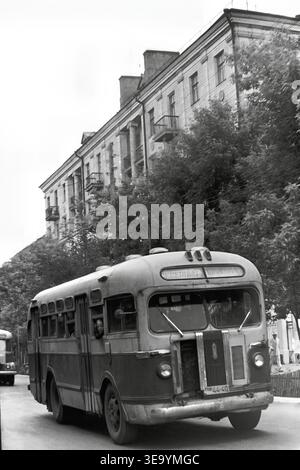 Sloviansk, 1956-1960, via Shevchenko. Questa straordinaria fotografia d'archivio storica offre una vista dettagliata ravvicinata di un classico autobus pubblico ZIS-155. Il robusto veicolo retrò, con i suoi ampi parabrezza e la griglia frontale in rilievo, domina la strada asfaltata. I passeggeri possono essere visti attraverso i finestrini laterali mentre l'autobus naviga lungo il viale alberato. Un grande edificio residenziale a più piani con colonne decorative e balconi forma uno sfondo elegante, che mostra la bellezza della tranquilla Donbas. Foto Stock