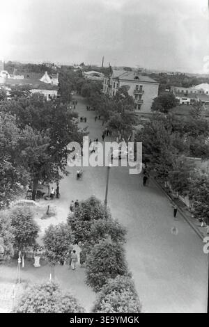 Sloviansk, 1956-1960, Piazza della Rivoluzione d'ottobre (ora Soborna). Questa accattivante fotografia d'archivio storica offre una vivace prospettiva elevata del traffico urbano centrale. Una classica autovettura di Pobeda e un camion pesante sono visti navigare lungo l'ampio viale asfaltato. Numerosi pedoni in abbigliamento vintage passeggiano lungo i marciapiedi ombreggiati. La vivace scena stradale è splendidamente incorniciata da fitti alberi maturi e dalla classica architettura residenziale sovietica, che cattura l'essenza della tranquilla Donbas. Foto Stock