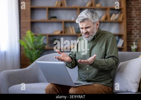 Uomo maturo con capelli grigi che esprime rabbia e confusione mentre guarda il suo laptop, seduto su un divano nel suo salotto, riscontrando problemi tecnologici o un errore di sistema Foto Stock