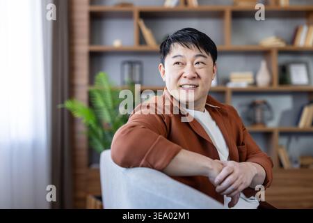 Uomo asiatico sorridente seduto comodamente in un ufficio o in un soggiorno, guardando lontano con cura con un'espressione felice, che rappresenta il relax, la fiducia e il comfort Foto Stock
