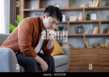 Uomo asiatico seduto su un divano nel suo salotto, guardando premuroso e preoccupato, lottando con lo stress emotivo, problemi di salute mentale, e sentendosi sopraffatto Foto Stock