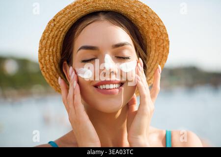 Donna che tocca il viso con entrambe le mani con la crema solare sulle guance. Giovane donna sorridente a occhi chiusi. Modello femminile che ama la routine della cura della pelle. Protez. Solare Foto Stock