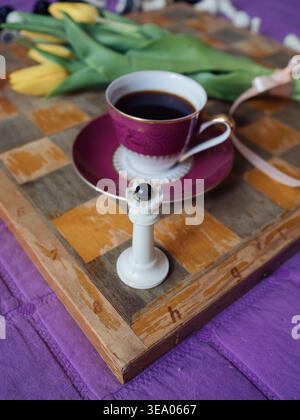 Regina degli scacchi bianca, tazza da caffè vintage e tulipani gialli su scacchiera, natura morta verticale mattutina accogliente Foto Stock