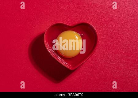 Tuorlo d'uovo di pollo crudo in un recipiente di ceramica a forma di cuore rosso su fondo rosso testurizzato. Fotografia gastronomica minimalista dall'alto per San Valentino o per la guarigione Foto Stock