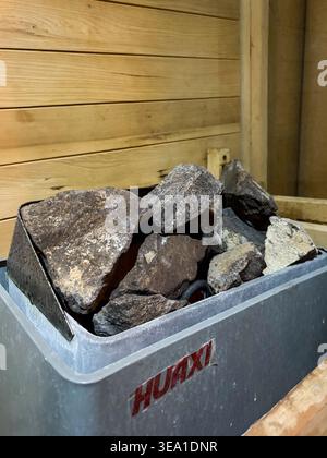 Pietre scure si trovano in un contenitore metallico all'interno di una sauna di legno. Le rocce ruvide poggiano in un vassoio poco profondo Pareti in legno circondano il contenitore del riscaldamento. Foto Stock
