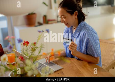 Una donna allegra siede a un tavolo di legno, fa shopping online con il suo laptop. Possiede una carta di credito, circondata da fiori e una bevanda rinfrescante. Foto Stock