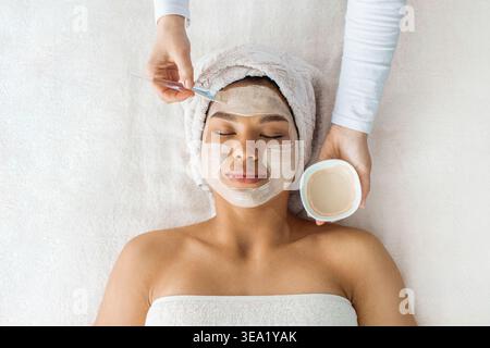 estetista che applica la maschera facciale in argilla bianca su una giovane donna nera alla spa Foto Stock