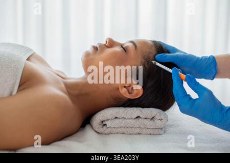 Donna nera che riceve un'iniezione facciale cosmetica nell'area del tempio nella clinica termale Foto Stock