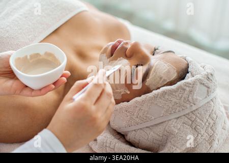 Estetista applicando maschera di argilla bianca con spatola su giovane donna nera Foto Stock