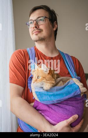Uomo che indossa una t-shirt arancione e occhiali che tiene in mano il suo amato gatto allo zenzero arancione comodamente accoccolato in un elegante portabicchieri in tessuto all'interno di una casa, mostrando il forte legame animale-umano domestico Foto Stock