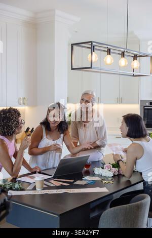 Donna anziana e tre donne adulte che si riuniscono a Kitchen Island, preparano carte con laptop e fiori Foto Stock