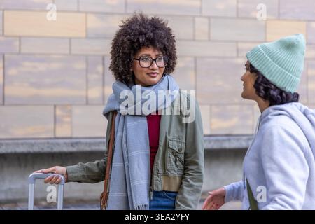 Due donne che parlano contro il muro di pietra su plaza, una che indossa una giacca di oliva che regge la valigia rotolante Foto Stock
