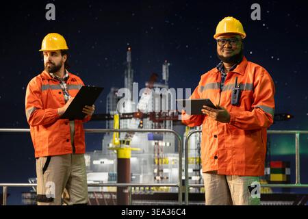 Ritratto di un team di piattaforme offshore sorridente che utilizza un tablet per controllare i sistemi sottomarini a tarda notte. Colleghi allegri che monitorano il rilevamento delle anomalie utilizzando il dispositivo per perforare derrick Foto Stock