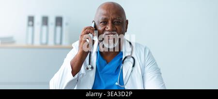 Medico in cappotto bianco effettuare una chiamata telefonica mentre si è seduti alla scrivania in Ufficio medico Foto Stock