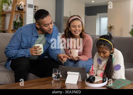 Famiglia con genitori e bambini seduti su un divano grigio, con tablet e scrittura su un notebook a spirale Foto Stock