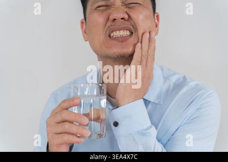 Primo piano di un uomo asiatico che tiene un bicchiere d'acqua mentre preme la guancia nel dolore, illustrando mal di denti, sensibilità dentale, infiammazione orale, e. Foto Stock