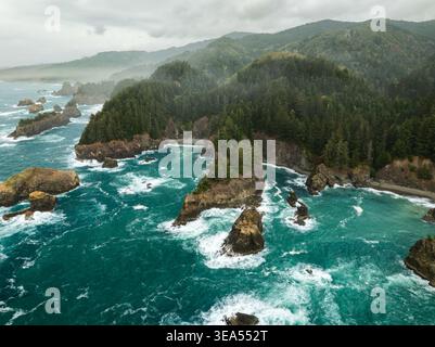 Vista aerea delle aspre montagne rocciose del mare e delle onde turchesi dell'oceano che si infrangono contro le fitte foreste sempreverdi sulla costa nebbiosa vicino alla costa dell'Oregon Foto Stock
