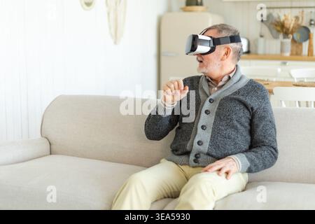 Un uomo anziano entusiasta indossando occhiali VR in realtà virtuale metaverse a casa. Nonno che tocca l'aria durante l'esperienza di realtà virtuale su casco per realtà virtuale. Videogame di simulazione ad alta tecnologia Foto Stock