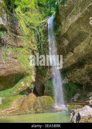 Cascata che scorre nella piscina limpida nella foresta verde con piccolo arcobaleno e luce del sole sulle rocce Foto Stock