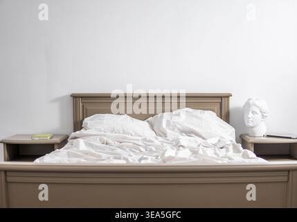 Letto non fatto in una camera minimalista con parete bianca e busto classico, simboleggia il riposo e la routine mattutina Foto Stock