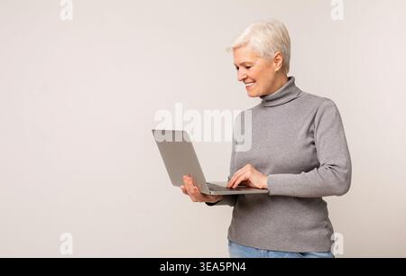 Donna anziana che utilizza un notebook mentre si trova su uno sfondo neutro in un ambiente interno luminoso Foto Stock