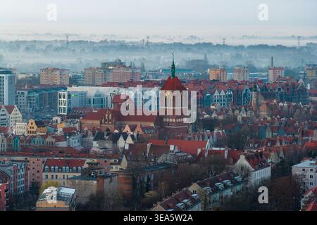 Vista aerea della storica chiesa di Santa Caterina e dei tradizionali edifici con tetti rossi sotto un cielo mattutino nebbioso a Danzica, nel Voivodato della Pomerania, P Foto Stock