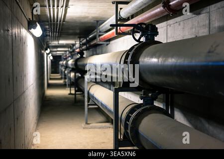 Un lungo corridoio poco illuminato in un impianto industriale caratterizzato da una serie di grandi tubi e valvole che corrono lungo le pareti e il soffitto Foto Stock