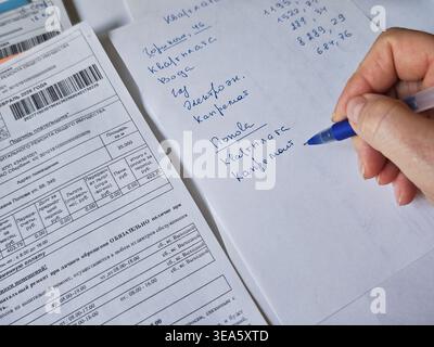 Kirov, Russia - 18 marzo 2026: La persona scrive le spese mensili e i pagamenti delle utenze mentre esamina le ricevute e i documenti finanziari su una scrivania Foto Stock
