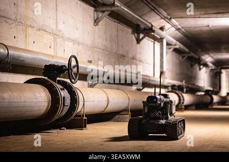 Un piccolo robot cingolato con antenne e telecamera è posizionato in un tunnel industriale buio accanto a tubi e valvole di grande diametro, il che suggerisce specifiche Foto Stock