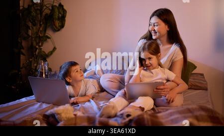 Mamma con bambini che usano tablet e smartphone a letto con una calda luce serale. Foto Stock