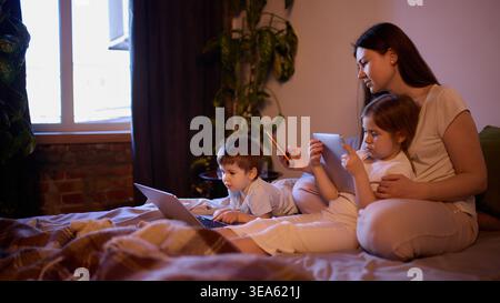 Mamma e bambini che utilizzano il laptop, il tablet e il telefono insieme sul letto, sotto la luce della sera. Foto Stock