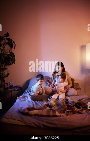 Mamma e bambini si rilassano sul letto utilizzando smartphone e laptop con una calda illuminazione serale. Foto Stock