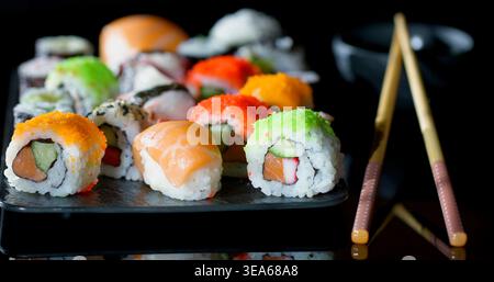 Molti tipi di sushi disposti su un piatto nero con bacchette e una piccola ciotola sullo sfondo Foto Stock