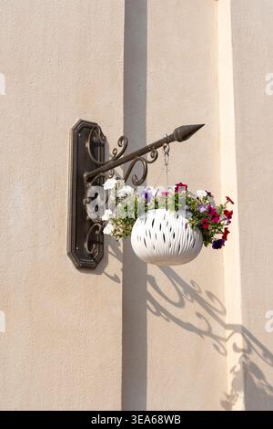 Vista verticale di una staffa decorativa in ferro battuto che regge una piantatrice sospesa bianca con fiori di petunia rosso, viola e bianco in fiore Foto Stock