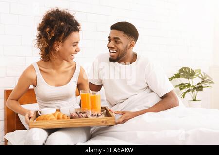 Coppia che gusta la colazione insieme nel letto con vassoio in legno di cibo e bevande in un ambiente accogliente Foto Stock