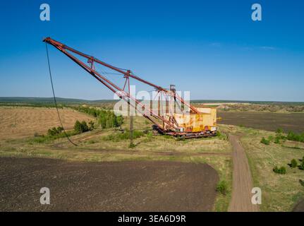 L'enorme escavatore dragline industriale poggia in un campo aperto, circondato da vegetazione sparsa e strade sterrate Foto Stock