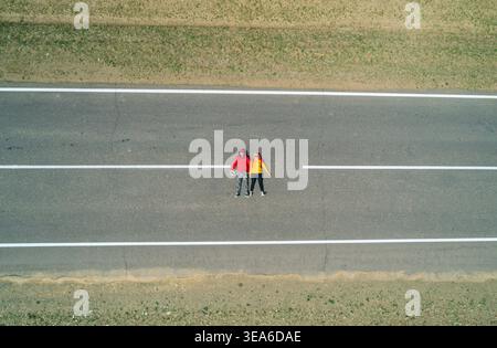 Vista aerea di una coppia di viaggiatori riposano su una strada vuota rettilinea, godendo di tranquillità con le mani dietro la testa Foto Stock