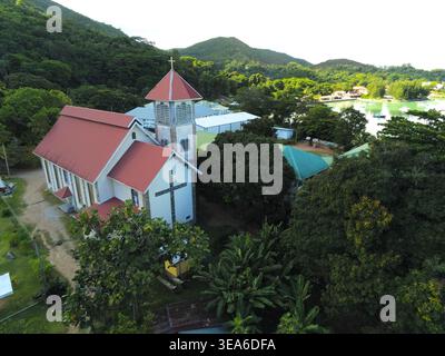Vista aerea della chiesa cattolica di Sant'Anna con il suo tetto rosso e il campanile circondato da lussureggiante vegetazione tropicale e colline a Baie Ste Anne, Praslin, se Foto Stock
