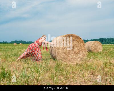 Una donna che indossa un abito a motivi rossi e occhiali da sole spinge una balla di fieno rotonda in un ampio campo verde e marrone. Foto Stock