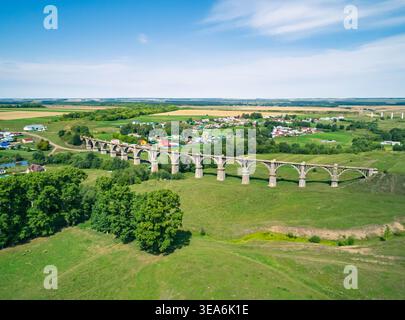 La vista aerea cattura lo storico ponte ferroviario ad arco che attraversa una vibrante valle verde accanto a un tranquillo villaggio rurale durante il giorno Foto Stock