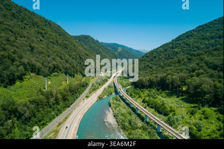 La vista dall'alto mostra un fiume tortuoso, un'autostrada trafficata e un imponente ponte ferroviario che attraversa una vibrante valle di montagna verde. Foto Stock