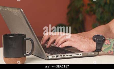 Persona che digita sul laptop con tazza da caffè e polso tatuato in un ambiente di lavoro domestico informale Foto Stock