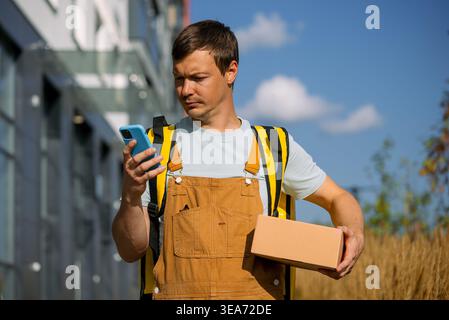 Addetto alle consegne che indossa tute e uno zaino termico utilizzando uno smartphone per la navigazione e la gestione degli ordini, trasportando un pacco su una strada urbana, rappresentando la logistica moderna e l'economia dei concerti Foto Stock