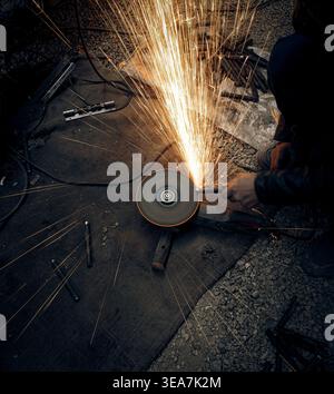 Scintille luminose volano mentre un lavoratore utilizza una smerigliatrice angolare in un'officina buia Foto Stock